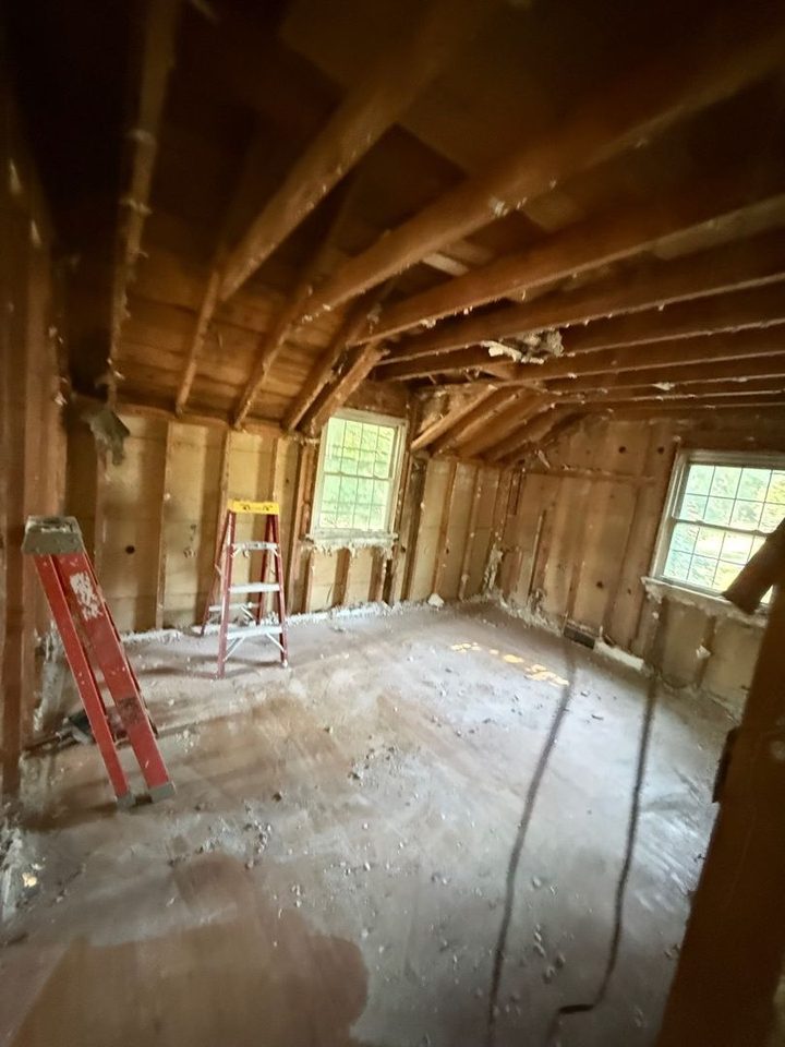 Attic room with walls and ceiling removed down to the wood framing, two windows and ladders on a bare floor